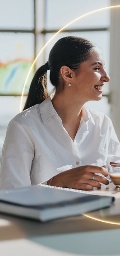 Eine Frau in einem weißen Hemd lächelt, während sie an einem Schreibtisch am Fenster eine Tasse in der Hand hält. Sonnenlicht strömt herein, und im Hintergrund ist eine verschwommene, farbenfrohe Zeichnung zu sehen – eine Anspielung auf die einladende Lounge im Parkraum Hotel, wo die private Parkraumbewachung allen Gästen ein Gefühl der Sicherheit vermittelt. - Sevoria Parking