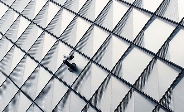 Eine Überwachungskamera, die an der Fassade eines modernen Gebäudes angebracht ist, deren geometrisches Muster aus sich überlappenden Glas- oder Metallpaneelen ein gitterartiges Erscheinungsbild erzeugt, eignet sich ideal für Anwendungen wie die private Parkraumbewachung oder ANPR-Systeme im Umfeld von Parkraumsystemen in Hotels. - Sevoria Parking