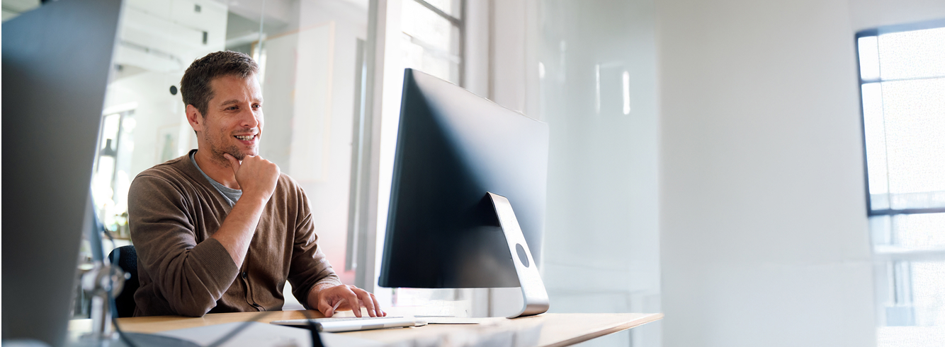 Ein Mann sitzt an einem Schreibtisch in einem hellen Büro, blickt auf einen großen Computerbildschirm und lächelt, während er mit der Hand am Kinn innovative Lösungen für das Parkraum-Hotelmanagement prüft und die Vorteile des schrankenlosen Parkens erkundet. - Sevoria Parking