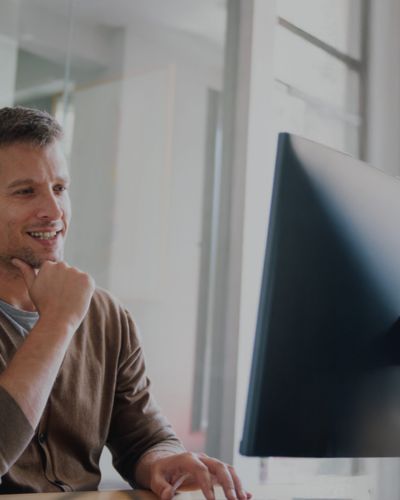 Ein Mann sitzt lächelnd an einem Schreibtisch und blickt auf einen großen Computerbildschirm in einem hellen Büro, wo er Lösungen für den Parkraumbereich im Einzelhandel verwaltet und sich mit den neuesten Technologien für barrierefreies Parken beschäftigt. - Sevoria Parking