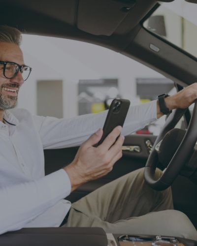 Ein Mann mit Brille und weißem Hemd sitzt auf dem Fahrersitz eines Autos, lächelt, während er auf sein Smartphone schaut – und genießt den Komfort des schrankenlosen Parkens, der durch die innovative ANPR-Parkraummanagement-Technologie ermöglicht wird. - Sevoria Parking