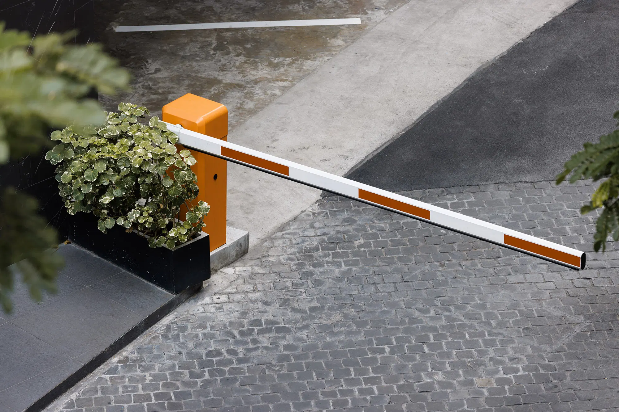 Am Eingang eines mit Kopfsteinpflaster gepflasterten Parkplatzes, neben einem Pflanzkübel mit grünen Blattpflanzen, ist ein orange-weiß gestreifter Parkplatzschrankenarm angehoben, was ein innovatives Parkraummanagement veranschaulicht, bei dem möglicherweise ANPR-Systeme für schrankenloses Parken zum Einsatz kommen. - Sevoria Parking