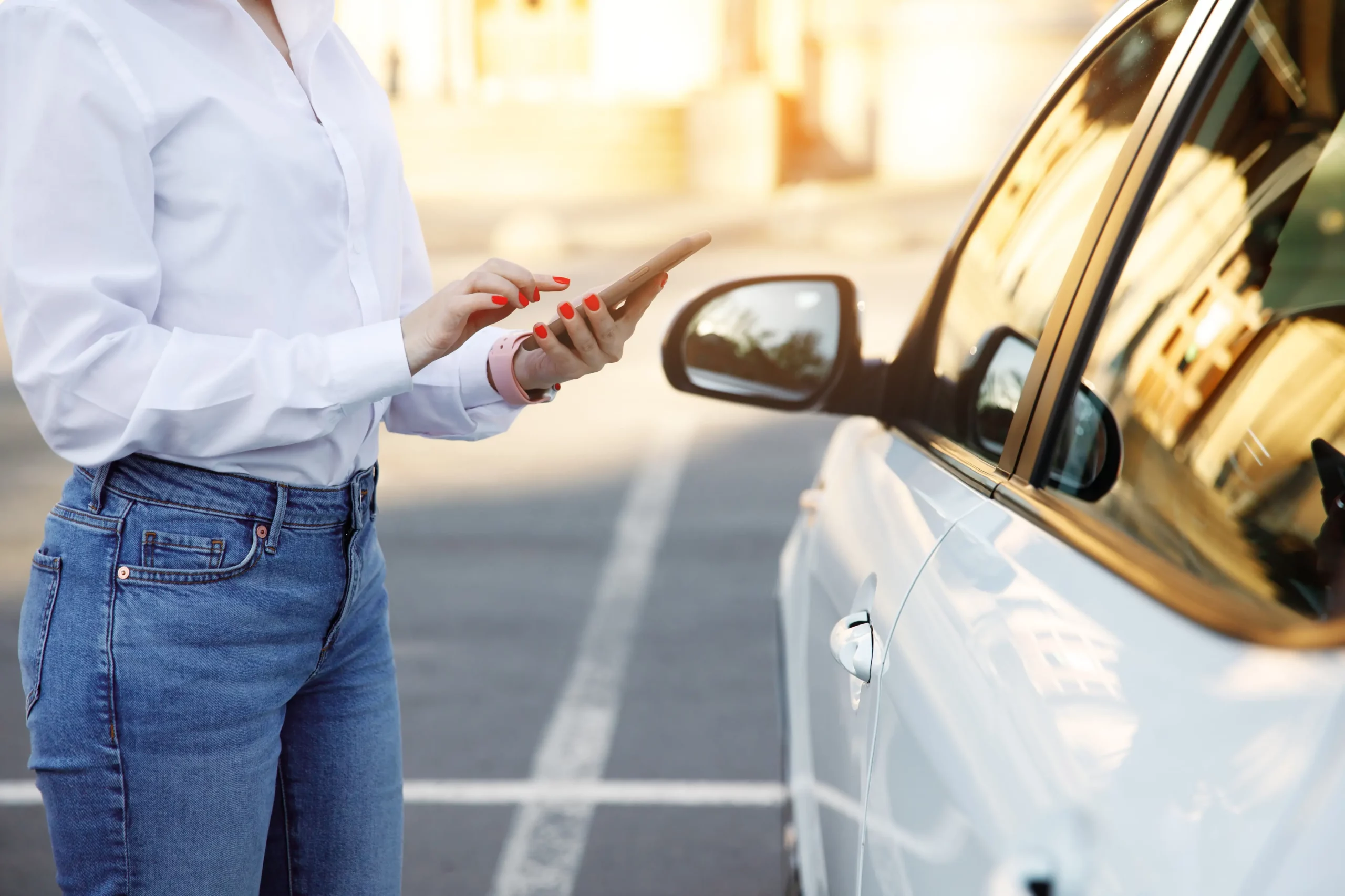 Eine Person in weißem Hemd und Jeans nutzt ihr Smartphone, während sie neben einem weißen Auto auf einem Parkplatz steht; möglicherweise informiert sie sich über Parkraummanagement-Optionen für Geschäfte in der Nähe oder verwaltet ihre Parkraum-Hotelreservierung. - Sevoria Parking