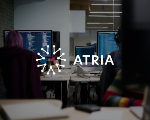 Zwei Personen arbeiten an Schreibtischen vor Computerbildschirmen, auf denen Programmcode zu sehen ist, und konzentrieren sich auf Lösungen für die Parkraumbewirtschaftung im Einzelhandel und im Hotelgewerbe. Über dem Bild sind das Wort „ATRIA“ und ein kreisförmiges Logo zu sehen, während der Hintergrund eine Büroumgebung mit Glaswänden zeigt – ein professionelles Umfeld, das sich der Innovation im Bereich der privaten Parkraumbewachung widmet. - Sevoria Parking