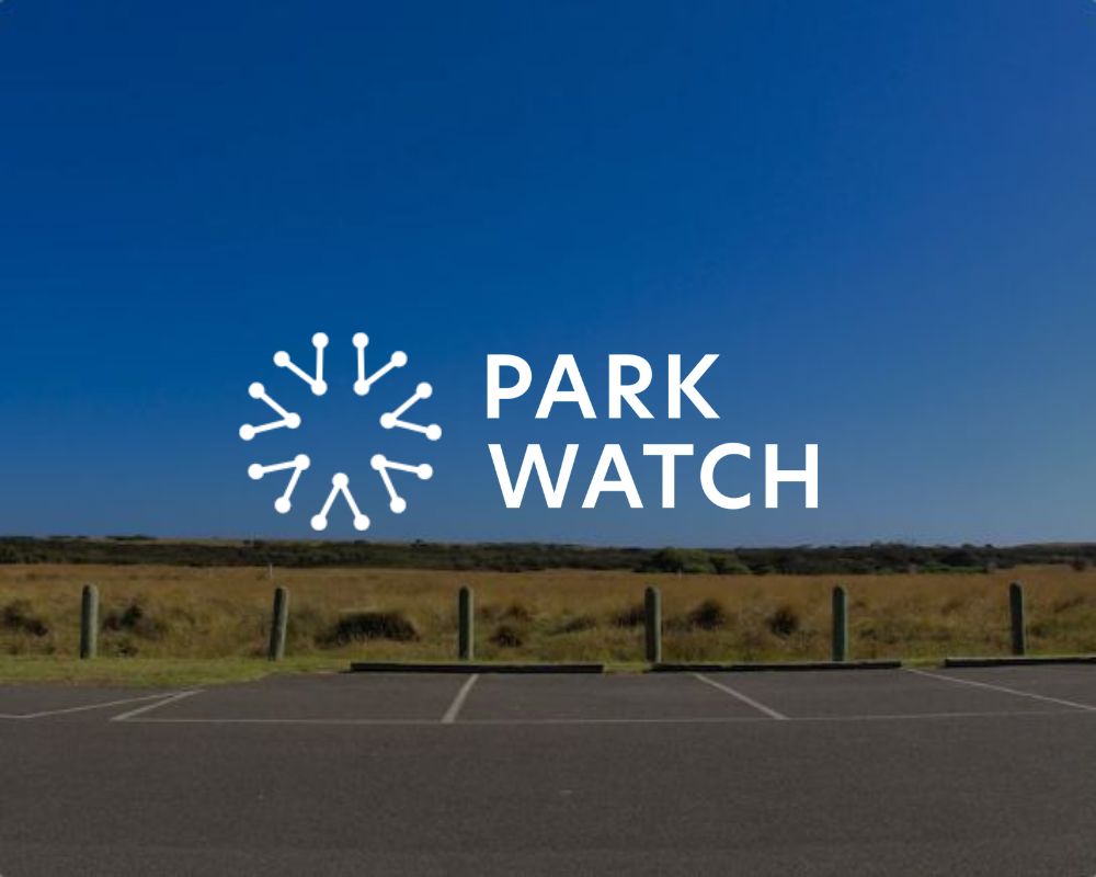 Ein leerer Parkplatz neben einer Wiese unter strahlend blauem Himmel, überlagert von einem runden weißen Logo und dem Schriftzug „PARK WATCH“ in fetter weißer Schrift, der die private Parkraumbewachung und fortschrittliche ANPR-Technologie hervorhebt – ideal für ein effizientes Parkraum-Hotelmanagement. - Sevoria Parking