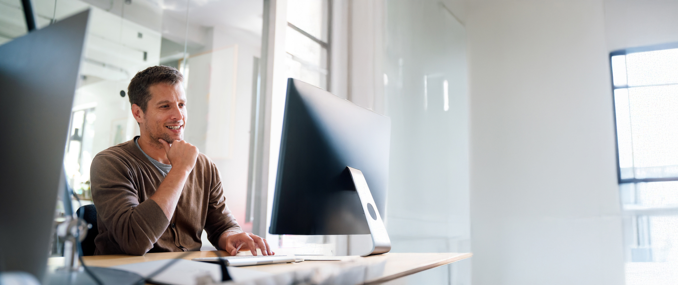 Ein Mann sitzt an einem Schreibtisch in einem hellen Büro und blickt lächelnd auf einen großen Computerbildschirm, während er sich die neuesten Lösungen für das Parkraummanagement ansieht, die ein effizientes schrankenloses Parken in einem stark frequentierten Parkraumhotel ermöglichen sollen. - Sevoria Parking