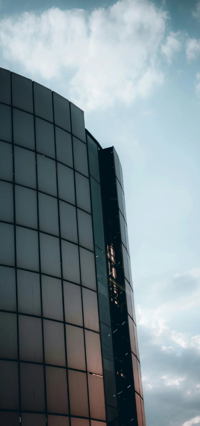 Modernes Bürogebäude aus Glas mit geschwungener Architektur, das das sanfte Licht des Himmels reflektiert. Vor dem Hintergrund eines teilweise bewölkten blauen Himmels wirkt das Gebäude hoch und schlank – ideal für hochwertige Einzelhandelsflächen, die einen zeitgemäßen Auftritt suchen. - Sevoria Parking