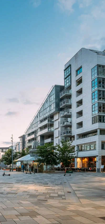 Moderne Wohnblocks mit großen Fenstern und Balkonen säumen eine breite, offene Fußgängerzone, die von lebhaften Einzelhandelsgeschäften gesäumt ist. Bei Sonnenuntergang sieht man auf dem Bürgersteig Sitzgelegenheiten im Freien, Bäume und Elektroroller, die eine lebhafte städtische Atmosphäre schaffen. - Sevoria Parking