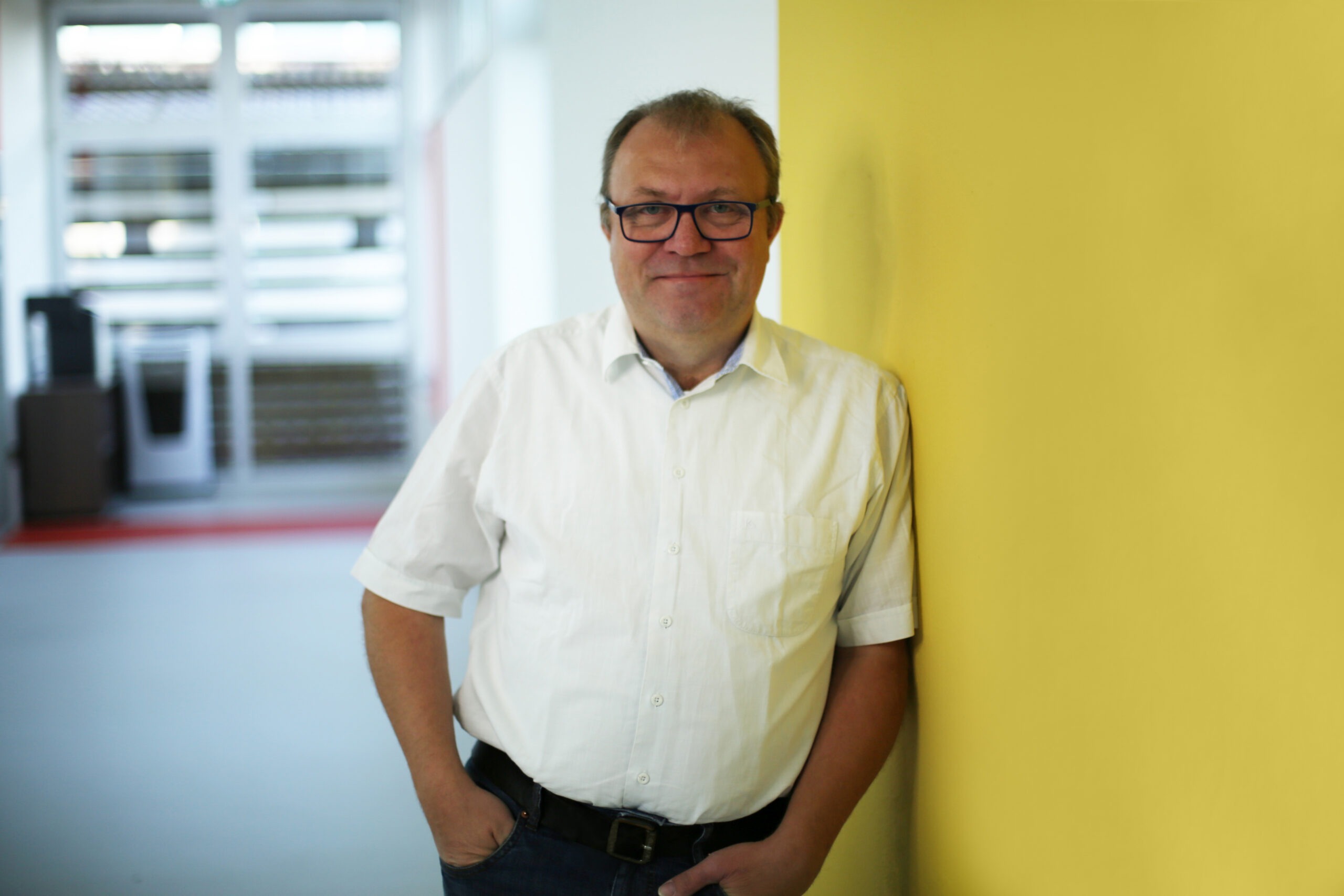 Ein Mann mit Brille und weißem Hemd steht lässig mit den Händen in den Taschen an einer gelben Wand in einem hell beleuchteten Innenraum und vermittelt damit die professionelle Atmosphäre, wie sie häufig im Bereich der Parkraumüberwachung und des Parkraummanagements anzutreffen ist. - Sevoria Parking